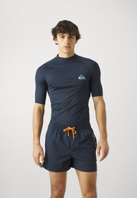 Granatowy rash guard z krótkim rękawem i wysokim kołnierzem, z jasnoniebieskim logo. W połączeniu z dopasowanymi granatowymi szortami kąpielowymi i pomarańczowym sznurkiem.