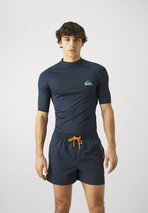 Jūras zila rash guard ar īsām piedurknēm un augstu apkakli, ar gaiši zilu logotipu. Saskaņots ar atbilstošām jūras zila peldbiksēm un oranžu saiti.