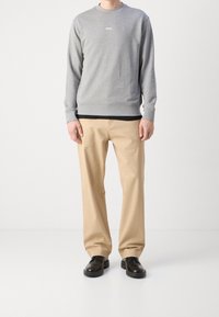Hombre vistiendo un sudadera gris de BOSS, pantalones beiges y zapatos de cuero negros, de pie contra un fondo blanco liso.