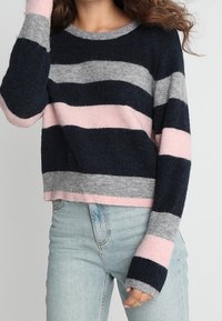 Žena v námořnickém, šedém a růžovém pruhovaném svetru s dlouhým rukávem, v kombinaci se světle modrými džíny, stojící s jednou rukou blízko obličeje.