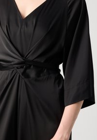 Robe noire avec un décolleté en V et un nœud torsadé à la taille, avec des manches trois-quarts et une texture de tissu lisse.