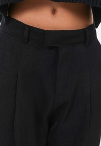 Pantalon noir avec un devant plat, une coupe droite et des plis visibles. Fabriqué dans un tissu lisse, il a une finition soignée et une taille.