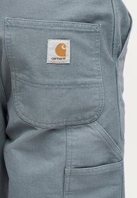 Λεπτομέρεια σε ανοιχτό γαλάζιο παντελόνι Carhartt με ραμμένες τσέπες και μια μικρή λευκή ετικέτα με πορτοκαλί λογότυπο Carhartt.