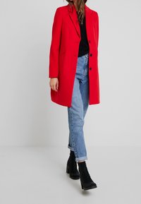 Frau, die einen roten, geknöpften Mantel, ein schwarzes Oberteil, hellblaue, hochgekrempelte Jeans und schwarze, hochhackige Stiefeletten trägt, steht vor einem schlichten Hintergrund.