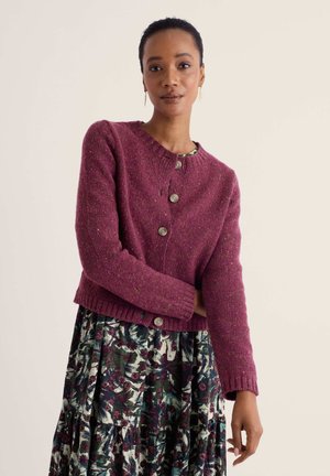 Femme aux cheveux courts portant des boucles d'oreilles en feuille d'or, un cardigan bordeaux boutonné et une jupe à motif floral sur un fond clair.