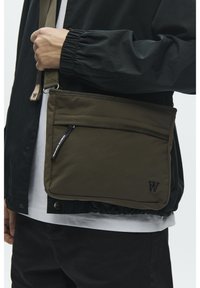 Oliiviroheline kandekott vastupidavast materjalist, millel on lukuga tasku, logoga detail ja reguleeritav rihm.