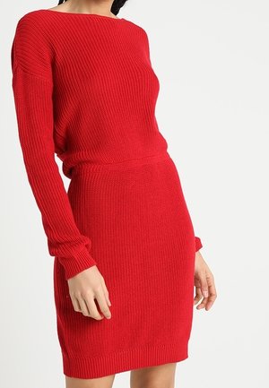Kobieta w dopasowanej, długorękawowej czerwonej sukience dzianinowej o prążkowanej fakturze, stojąca na tle jednolitego jasnego tła.