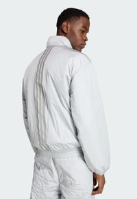 Chaqueta de plumón gris claro con tres franjas verticales en la parte de atrás, cintura elástica y puños de canalé, confeccionada en un material brillante y texturizado.