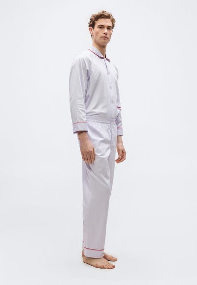 Άνδρας μοντέλο στέκεται ξυπόλητος φορώντας ανοιχτό γκρι πιτζάμα με κόκκινες ραφές στα μανίκια, το γιακά, την τσέπη και τα τελειώματα των παντελονιών, μπροστά σε απλό φόντο.