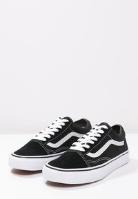 Paire de baskets basses noir et blanc avec lacets blancs et bande latérale blanche ondulée sur une surface blanche.
