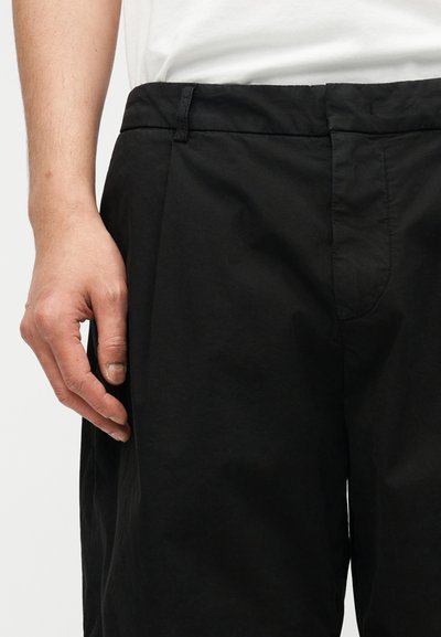 Αριστερό χέρι ακουμπισμένο δίπλα στο ισχίο, φοράει μαύρο παντελόνι και άσπρο πουκάμισο, με φόντο απλό ανοιχτόχρωμο.