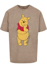 Beige oversized t-skjorte med en tegneserie-bjørn som har en gul kropp og rød skjorte, stående med den ene hånden på haken.