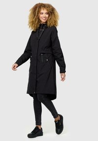 Abrigo negro largo con alto cuello, cordones ajustables y bolsillos laterales. Presenta una tela texturizada y una silueta elegante. Combinado con jeans oscuros y zapatillas.