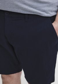 Marineblaue Shorts aus leichtem Stoff mit glatter Textur, ausgestattet mit einem Knopfverschluss, Gürtelschlaufen und Seitentaschen.