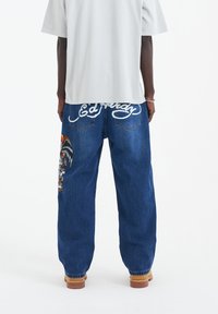 Niebieskie jeansy z denimu o luźnym kroju. Z tyłu biały haftowany napis oraz kolorowy wzór graficzny na lewej nogawce.