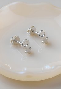 Silberne Steckerohrringe mit einem Design des weiblichen Symbols, polierte Oberfläche und einer runden Akzentuierung. Auf einem glatten, hellen Teller platziert.