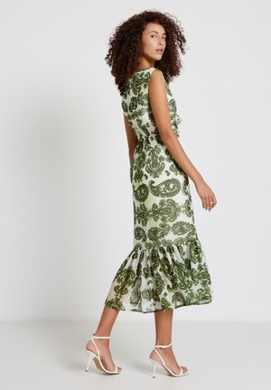 Femme aux cheveux bouclés portant une robe midi blanche sans manches avec des motifs baroques verts et des sandales blanches à talons hauts, debout de côté.