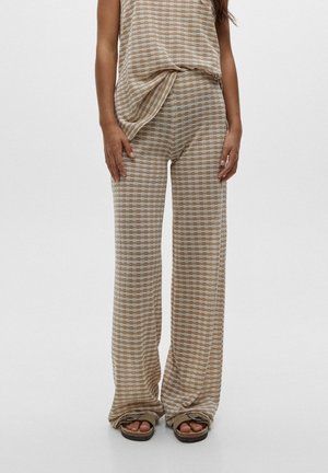 Femme portant un pantalon large beige et blanc à motifs et un haut sans manches assorti avec des sandales marron sur un fond blanc uni.