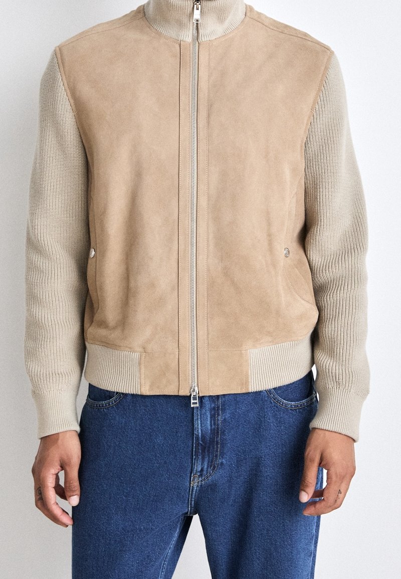 Veste beige clair zippée avec manches tricotées et poignets côtelés portée avec un jean en denim bleu, vue du torse jusqu'aux mains.