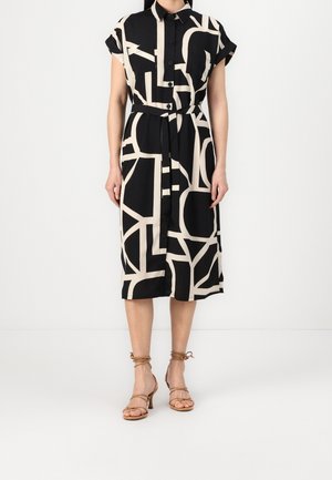 Mujer con un vestido midi abotonado de patrón geométrico en negro y beige, con mangas cortas y cinturón anudado en la cintura, combinado con tacones con tiras en color marrón claro.