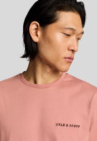 Camiseta de color rosa claro con cuello redondo, que presenta un logo bordado en negro en el lado izquierdo del pecho. Material de algodón suave, corte medio.
