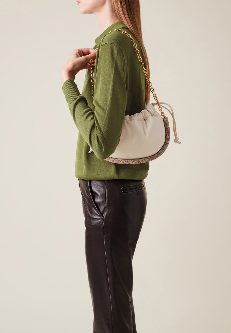 Cremfarbene Leder-Schultertasche mit strukturiertem braunem Boden, ausgestattet mit einer Kettenriemen und einem gerafften Design. Getragen von einem Model mit einem grünen Pullover.