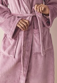 Lyserød tekstureret badekåbe med diamantmønster, brede ærmer, et bundet bælte og to forlommer, lavet af blødt, fluffy materiale.