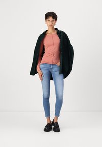 T-shirt rosa chiaro a maniche lunghe, abbinata a una camicia oversized verde scuro con bottoni. Jeans slim-fit blu e scarpe nere completano il look.