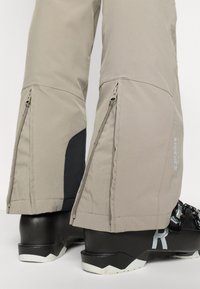 Bež hlače za smučanje z bočnimi zadrgami, opremljene z črnim poudarkom. Oblikovane tako, da se lepo prilegajo čez smučarske čevlje z rahlim razširjenjem na robu.