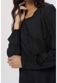 Femme portant une chemise noire à manches longues boutonnée, la main gauche touchant son cou et le bras droit croisé sur son torse.