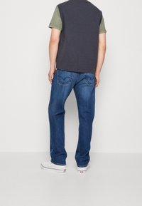 Laivastonsininen hihaton neule oliivinvärisen t-paidan päällä, yhdistetty sinisiin farkkuihin ja valkoisiin tennareihin. Näkyvissä ompeleet ja taskujen yksityiskohdat.