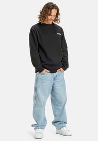 Sudadera de algodón negra con escote redondeado y pequeño logo. Combinada con unos vaqueros holgados de color azul claro y zapatillas blancas, creando un look casual.