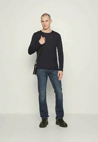 Camisa de manga longa azul-marinho combinada com jeans azuis. O modelo carrega uma bolsa preta de ombro e usa tênis escuros. Design simples e casual.