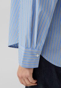 Himmelblaues Hemd mit vertikalen weißen und dunkelblauen Streifen. Verfügt über kleine eingeprägte silberne Akzente, Manschette mit Knöpfen und eine weiche Stofftextur.