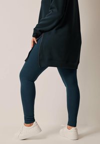 Leggings en verde azulado oscuro con una textura suave, combinados con un sudadera negra larga con cierre lateral y ajuste holgado, llevados con zapatillas blancas.