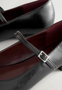 Black leather Mary Jane shoes with a silver buckle and dark red interior lining, shown in close-up detail.