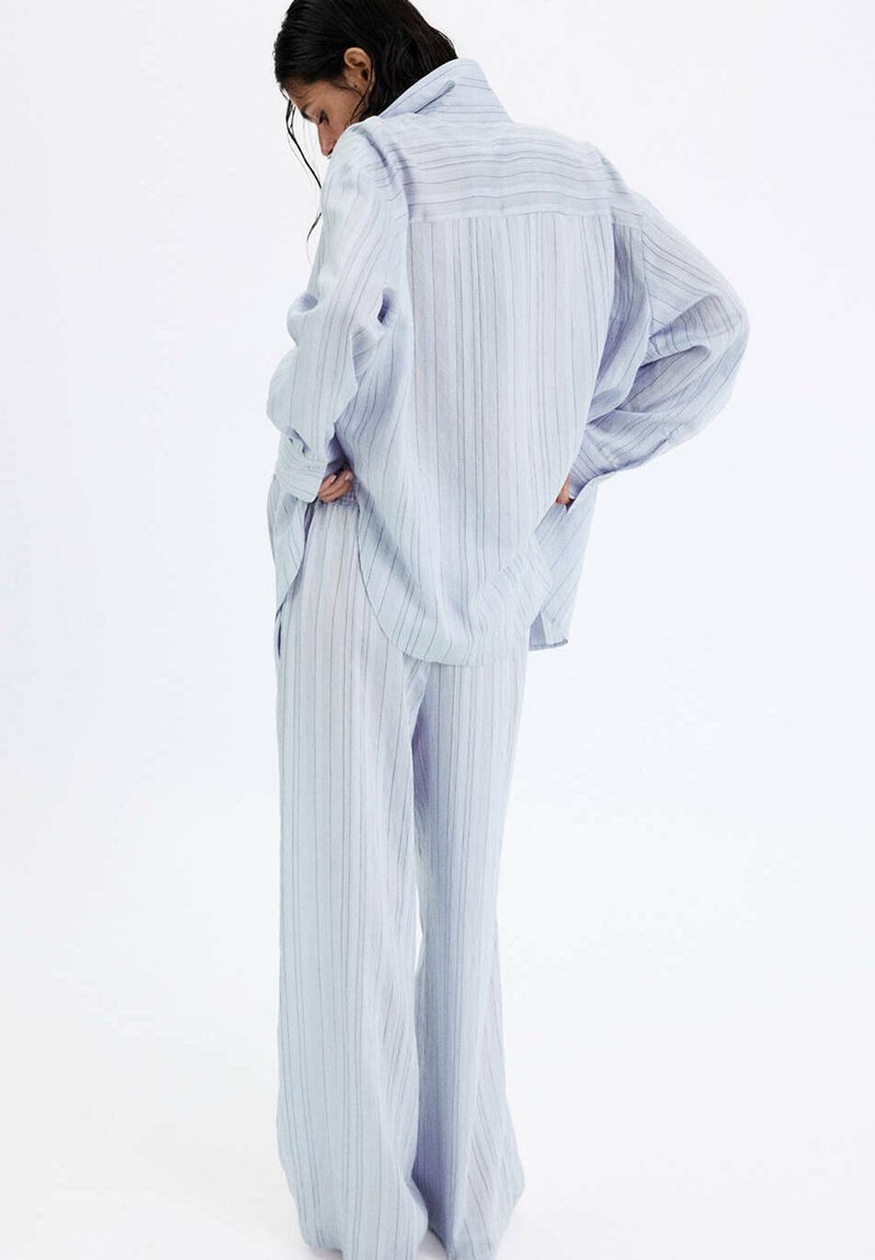 Femme aux cheveux foncés portant une chemise ample rayée bleu clair et un pantalon large assorti, debout les mains dans les poches, de dos.