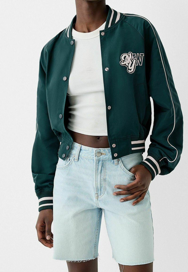 Personne portant une veste universitaire vert foncé avec des rayures blanches, un crop top blanc et un short en jean bleu clair avec des ourlets effilochés.