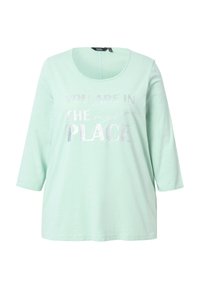 Mentolově zelené tričko s dlouhým rukávem a zaobleným výstřihem. Obsahuje stříbrný metalický potisk s textem: "JSTE NA SPRÁVNÉM MÍSTĚ." Měkká textura.