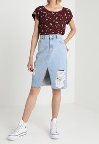 Rotes, blumengemustertes Kurzarmoberteil kombiniert mit einem hellblauen Denim-Bleistiftrock mit einem Schlitz vorne und ausgefransten Details. Weiße High-Top-Sneaker.