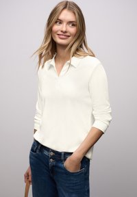 Polo bianca a maniche lunghe con collo a zip, abbinata a jeans di denim blu. Il tessuto è liscio con una leggera lucentezza.