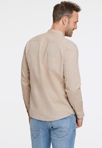 Lichtbeige, langärmelige shirt met een subtiele textuur, met knopen bij de manchetten en een afgeronde zoom, gedragen met lichtblauwe jeans vanuit een achteraanzicht.