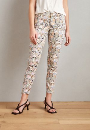 Kvinde iført slanke ankellange bukser med et pastel-paisley mønster, hvid bluse og sorte remsandaletter med hæl.