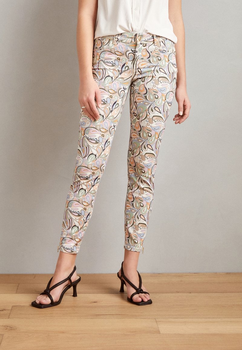 Femme portant un pantalon slim longueur cheville avec un motif cachemire pastel, un chemisier blanc et des sandales à talons noires à brides.