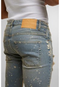 Izgubljene plave traperice imaju izblijedjeli izgled s oštećenjima i prskanom bijelom bojom, a na struku se nalazi smeđi kožni uložak s utisnutim tekstom.