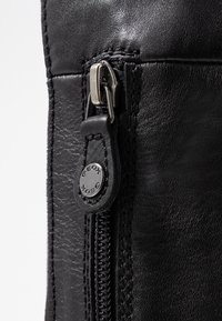 Detalle de un bolso de cuero negro con una cremallera metálica con tirador, mostrando una superficie texturizada y costuras en contraste a lo largo de la costura.