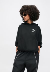 Μαύρο φούτερ με κουκούλα, μπροστινή τσέπη και κυκλικό λογότυπο. Συνδυάζεται με μαύρες φόρμες με λευκές ρίγες στο πλάι.