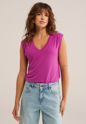 Femme aux cheveux mi-longs bouclés portant un haut sans manches magenta à col en V et un jean taille haute bleu clair, sur un fond neutre.