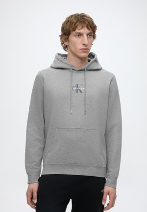 Młody mężczyzna w jasnoszarej bluzie z kapturem Calvin Klein z przednią kieszenią i sznurkami, połączonej z czarnymi spodniami, stojący na białym tle.