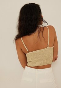 Woman with long dark hair wearing a beige textured crop top with thin straps and white high-waisted pants, facing away against a plain background.